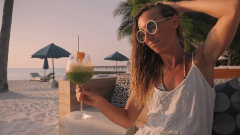 Woman having cocktail at sunset on tropical beach. Female having exotic drink in luxury resort by the beach