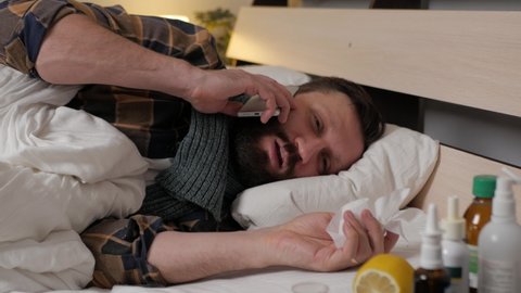 A sick man is lying in bed home, talking phone and wiping his nose with handkerchief. The man is upset and has difficulty communicating, his throat hurts. Season of colds, fever, infection, flu.