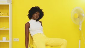 Young black woman, stylish model, is sitting on yellow chair next to a fan, chilling out and looking at camera confidently, smiling and posing, creative portrait video, Slow motion.