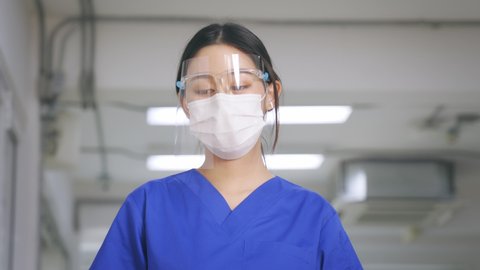 Young Asian nurse wearing uniform and rubbing eyes removing face shield while wearing surgical mask looking distraught and sad while taking break after hard work in hospital