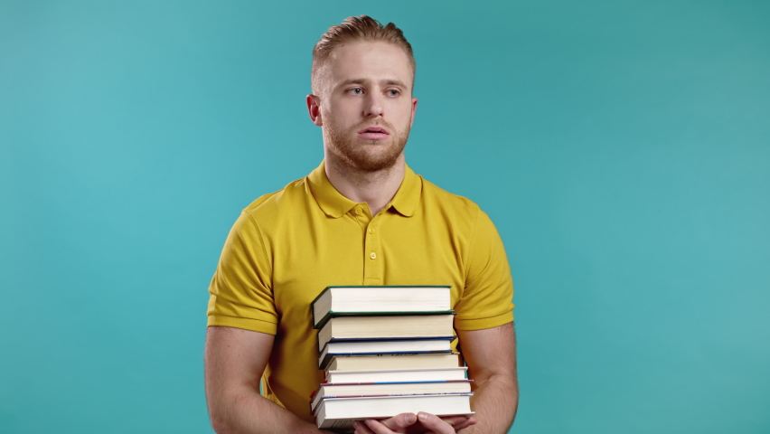 European student is dissatisfied with amount of homework and books on blue background. Man sighs and rolls his eyes in displeasure, he is annoyed, discouraged frustrated by studies. | Shutterstock HD Video #1068437645