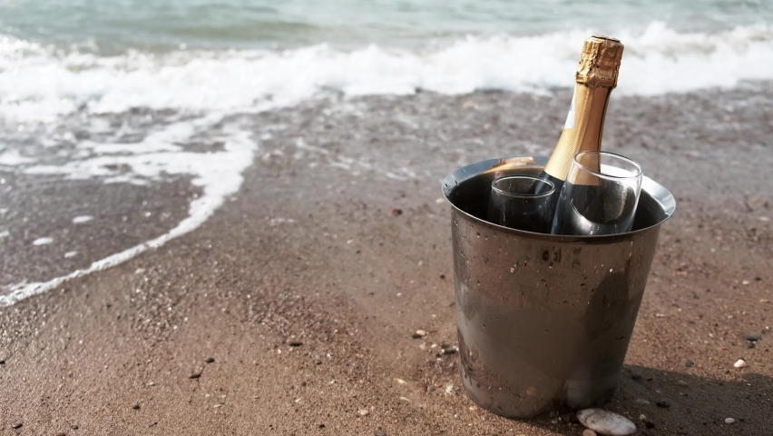 Ice bucket with champagne bottle and two glasses in sand washed by sea water. Waves from foaming ocean gently coming around. Romantic getaway, honeymoon, vacation or anniversary at luxury destination.