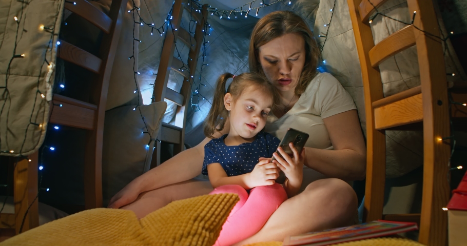 Cute baby daughter has fun with her mom enjoying using her smartphone showing the game on a modern gadget mobile phone looking at the mobile screen while sitting in a tent at home