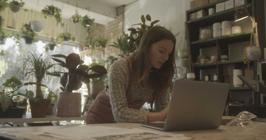 Young adult Female, small business owner working on laptop in store and speaking to client on phone | Shutterstock HD Video #1068440027