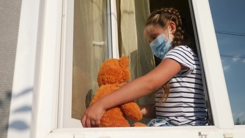 bored kid in medical mask in home quarantine coronavirus covid 19 sitting by the window. child with a toy teddy bear in protective mask looking out the window. coronavirus epidemic prevention concept