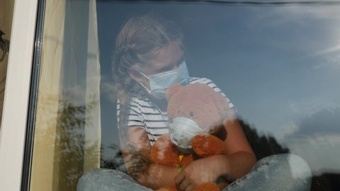 bored kid in medical mask in home quarantine coronavirus sitting by the window. child with a toy teddy bear in protective masks looking out covid 19 the window. coronavirus epidemic prevention concept