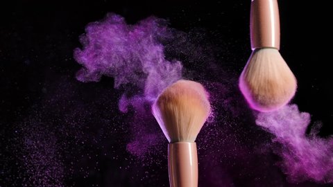 From the touch of two makeup brushes, colorful glittering powder or blush particles float in the air. Bright creative makeup, concept on black background in pink neon light. Slow motion. Close up.