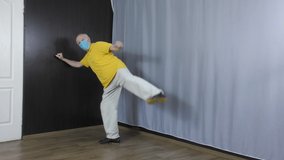 In a medical mask, an old man athlete makes a leg lift to the side near a white background