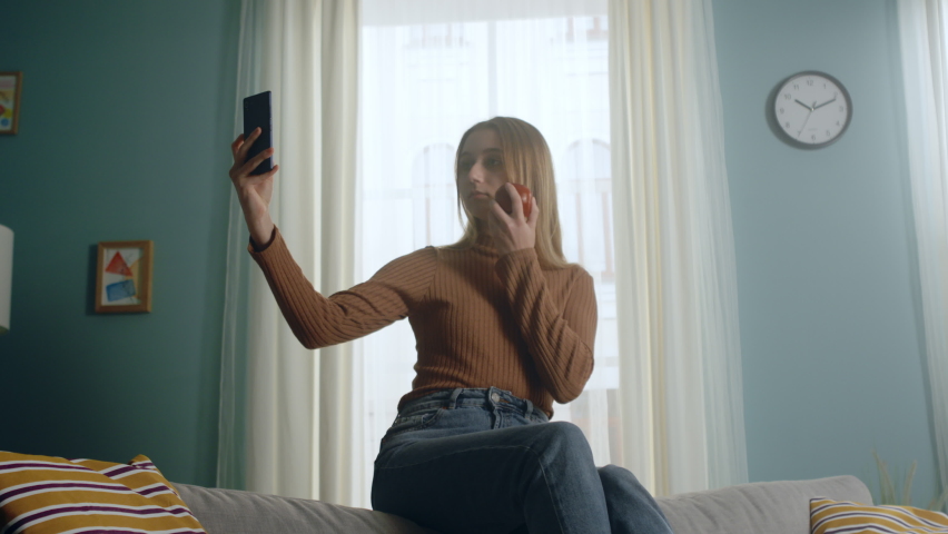Young woman in blue jeans and brown sweater sits on back of sofa on background of window, holding red apple and taking selfie. Average plan.