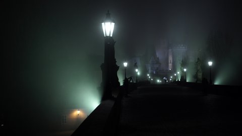 
lit lamps on Charles Bridge and illuminated stone statues and silhouettes of pedestrians at night and in the background the bridge tower in the fog