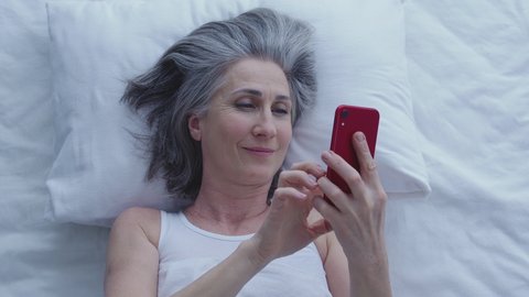 Happy greyhaired woman chatting online in bed using dating application, internet