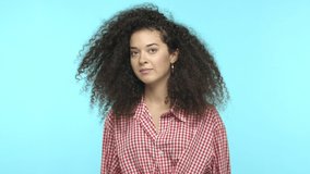 Slow motion of awkward young woman with curly hair, raising eyebrows shocked, cover eyes from embarrassing scene, unwilling to look, standing over blue background