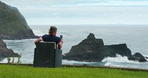 Back view of a man sitting by the ocean while using his cell phone. Caucasian male traveler texting in the picturesque area of the peninsula. High quality 4k footage