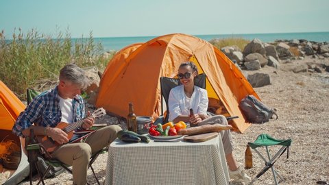 couple having fun in seaside camping at sunny day. handsome gray-haired man sitting in camp chair and playing on ukulele funny song for attractive joyful girlfriend