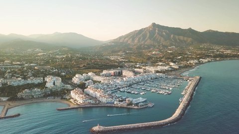 Cinematic aerial perspective of Puerto Banus Bay, Marbella. Beautiful golden hour with warm light reflection on the buildings . Boats leaving the port. La Concha mountain in background. Drone forward