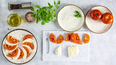 Making of Caprese salad with sliced fresh tomatoes, mozzarella cheese and basil served on a white plate on light gray table surface, top view. Traditional Italian food. Stop motion animation