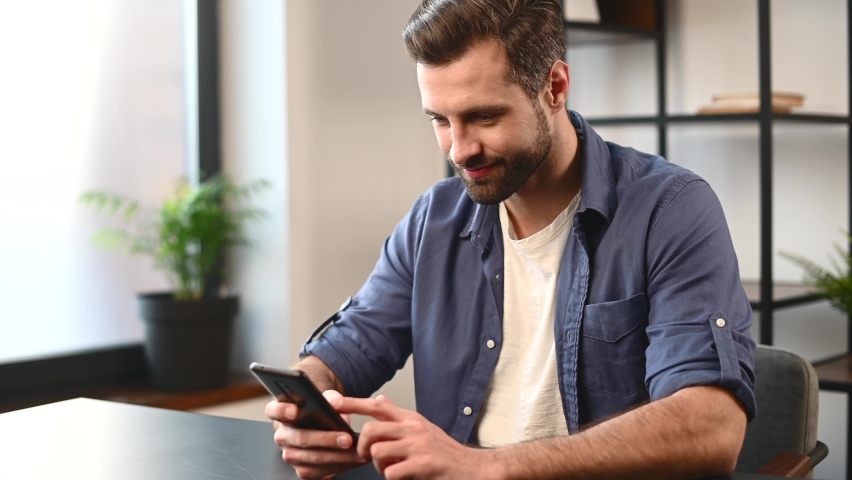 A handsome bearded guy spends leisure with a smartphone indoor, a man is scrolling news feed, watching funny videos and laughs sitting on the desk, chatting in networks or dating sites | Shutterstock HD Video #1068471107