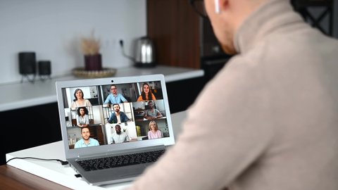 Back view man with earphones greeting diverse team on the laptop screen by video call, sits in the kitchen at home, new level of the workplace, telecommunication, online meeting with multiracial team