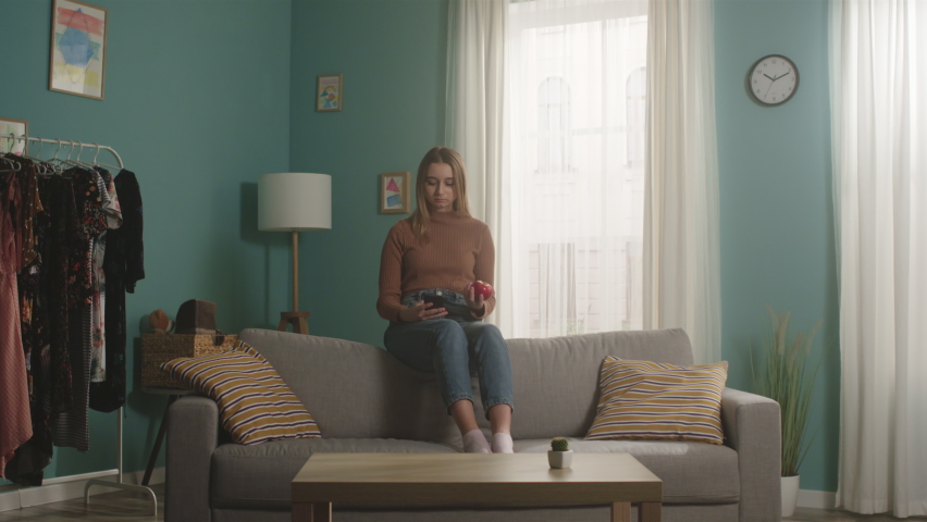 Young woman in blue jeans and a brown sweater sits on the back of a sofa, holds a red apple and takes a selfie. Long shot
