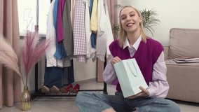 A beautiful young blonde woman sitting on the floor takes out a gift from a paper bag of pink sneakers. A female fashion stylist. 4k video.