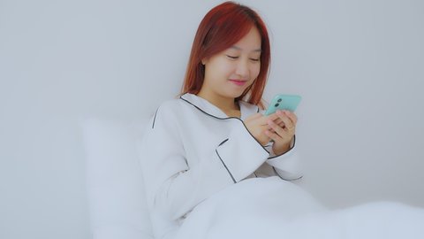 A beautiful asian woman red hair dye color in white pajamas on bed in bedroom. She play social media on mobile phone in the morning on holiday. Relax, smile and laugh good mood. Lifestyle Concept