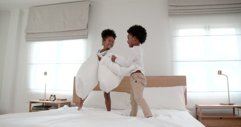 A little adorable African American boy and girl having a pillow fight together on bed in bedroom. A cute small black kids siblings having fun laughing playing funny active game. Family concept.