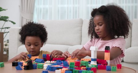 An older African American sister helps her little brother making a train and the city with a colorful Lego bricks in living room. Two cute african children siblings playing building block together.