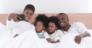 Happy African American family lying on bed in bedroom and making video call to talking with their cousin. Young parent using smartphone to record video with their adorable kids at home.