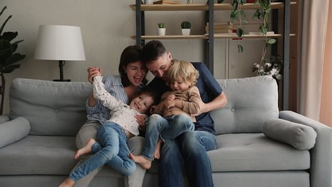 Happy loving parents sitting on cozy sofa, playing with small adorable preschool kids siblings at home. Smiling positive family enjoying spending weekend leisure time together, having fun indoors.