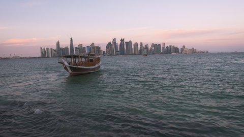 Doha, Qatar- January 16 2021:Doha skyline from the corniche promenade sunset zooming in shot showing dhow with Qatar flags in the Arabic gulf  in foreground and clouds in the sky in background