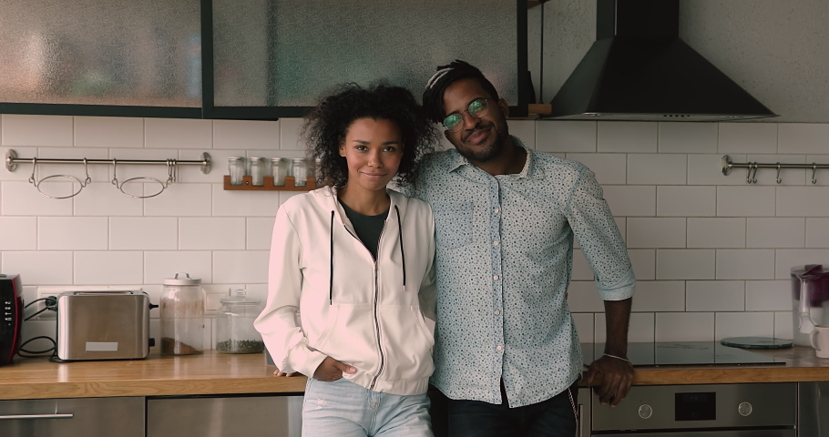 Happy young family african wife, husband with dreadlocks pleased good-looking 35s couple standing in domestic modern comfort remodelled kitchen smiling looking at camera. Homeowners portrait concept | Shutterstock HD Video #1068492926