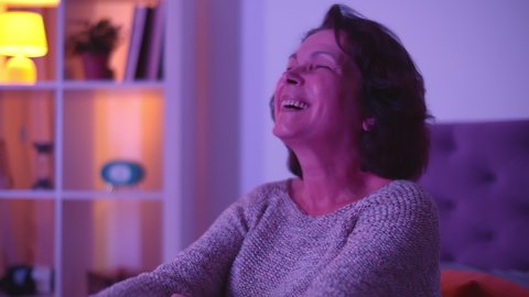 Senior woman laughing while watching tv at home in evening. Portrait of happy retired female smiling watching comedy movie relaxing in living room late at night