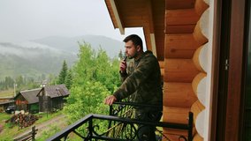 Handsome bearded man stands on the balcony of a country apartment in the mountains on a rainy day and smokes an electronic cigarette.