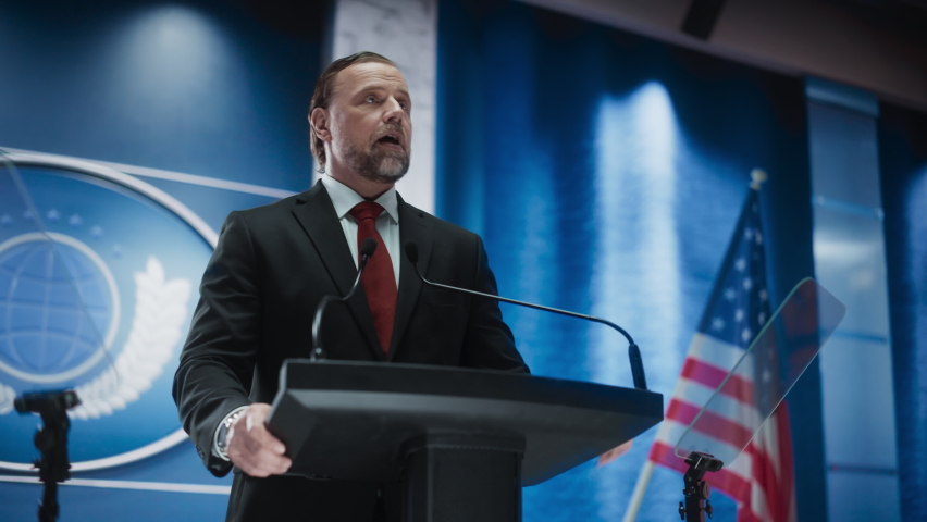 Portrait of Organization Representative Speaking at a Press Conference in Government Building. Press Officer Delivering a Speech at a Summit. Minister at Congress. Backdrop with American Flags. Royalty-Free Stock Footage #1068497879