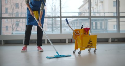 Uniformed cleaner wiping floor using mop and cart in business center. Cropped shot of janitor in apron and gloves cleaning floor in corridor of large office building