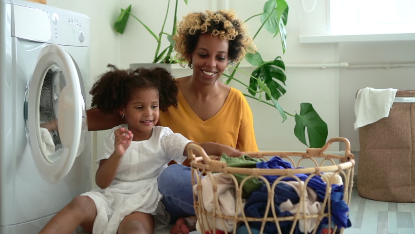 Happy family of mom and daughter are doing household chores in laundry at home spbd. African American woman and little girl put dirty clothes in washing machine and talk with smiles while sitting on Royalty-Free Stock Footage #1068502199