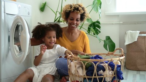 Happy family of mom and daughter are doing household chores in laundry at home spbd. African American woman and little girl put dirty clothes in washing machine and talk with smiles while sitting on
