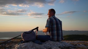 man drinks a hot drink from thermos bottle at evening on a mountain hike travel peak Spbd. A tourist admires the beautiful view of sunset. Hiking and sit on rock. backpack and trekkig poles concept