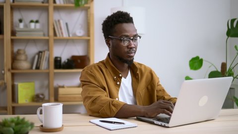African man in glasses works at home background of white interior Spbas. Serious male black-skinned model typing text on computer at table with cup of coffee and notebook. Green house plant in