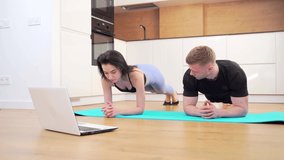  young active couple in sportswear doing planks on the floor in front of laptop while watching online workout. man and woman sports at home. family team does physical fitness exercises together