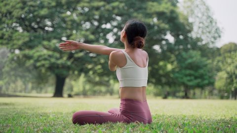Asian young woman sitting on the grass in the lotus position and raising hands up outside in city park with the big trees background. Rearview of female practicing yoga outdoors on a sunny day.