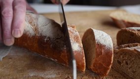 Cut crusty baguette into slices. Hands slice traditional french  baguette on wooden cutting board. Close-up slow motion movie with sound.