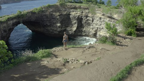 Aerial moving forward and panning a natural rock arch formation land bridge spanning a tropical bay and coastline with a woman admiring the view - Nusa Penida and Lombok, Bali