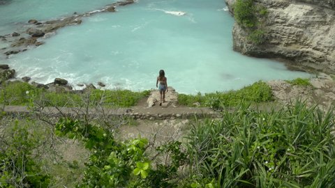 Aerial rising above a woman as she approaches a cyan ocean cove with steep rocky cliffs, lapping waves, and a tilt up to the larger ocean background stretching to the horizon - Nusa Penida, Bali