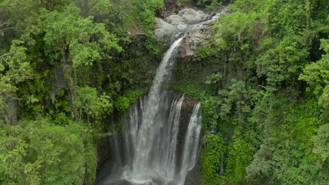 Aerial moving down into a tropical gorge with full view of a rushing waterfall surrounded by jagged cliff walls covered in dense foliage with cool mist rising from the base of the falls - Nusa Penida 