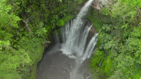 Aerial tilt up from a plunge basin to a rushing waterfall surrounded by jagged cliff walls covered in dense tropical foliage with cool mist clinging to the treetops - Nusa Penida and Lombok, Bali