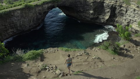 Aerial moving forward with a dramatic tilt down into a tropical circular bay with crashing waves, a natural rock arch bridge formation, and a woman admiring the view - Nusa Penida and Lombok, Bali