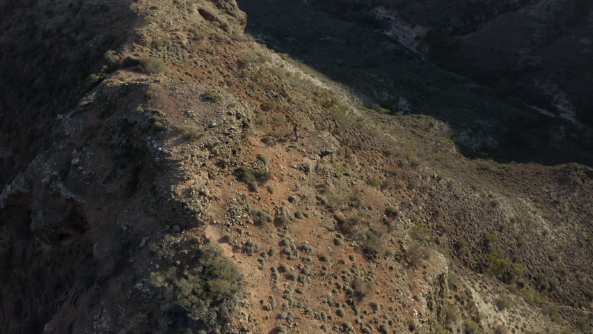 Aerial circling a woman hiker standing on a rocky ledge surrounded by remote, dry, desert hills with brush, rocks, bright sunlight, and blue sky - Exmouth, Australia