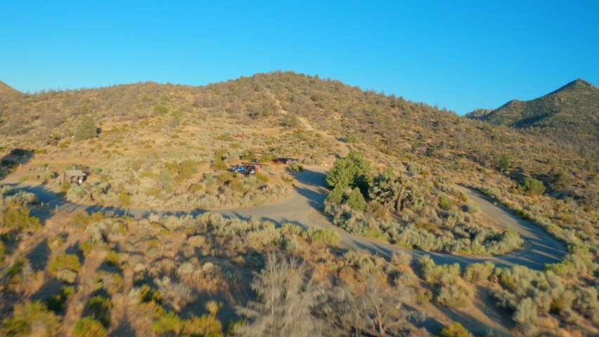Aerial moving forward over dry brush and towards rolling hills under a bright clear blue sky, with roads and a few cars parked along the way - Walker Pass, California