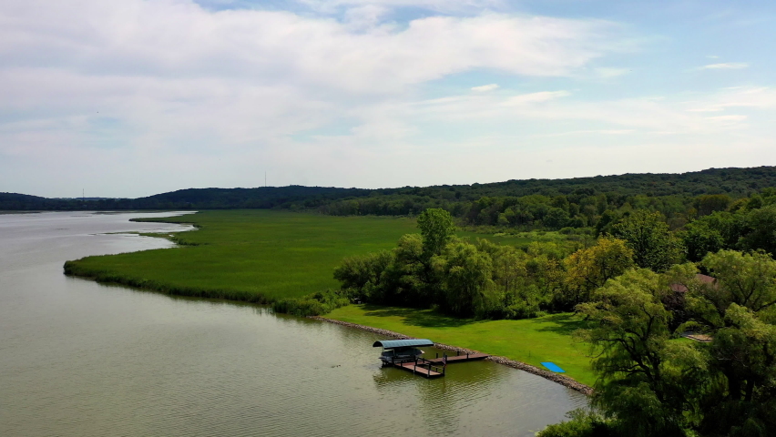 Aerial tilt down shot of green landscape by lake against cloudy sky, drone flying forward over grassland on sunny day - Lake Como, Wisconsin